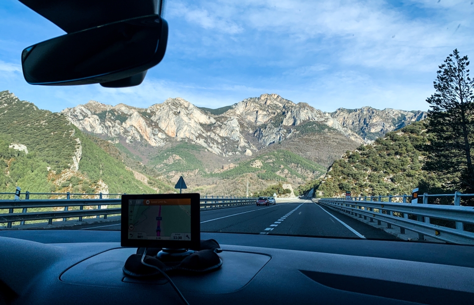 View from the front window of a car, GPS sits on the dashboard. Behind it a blue sky, scarcely clouded with a sedimentary mountain range. It has stripes of colors from green to grey to brown to beige.